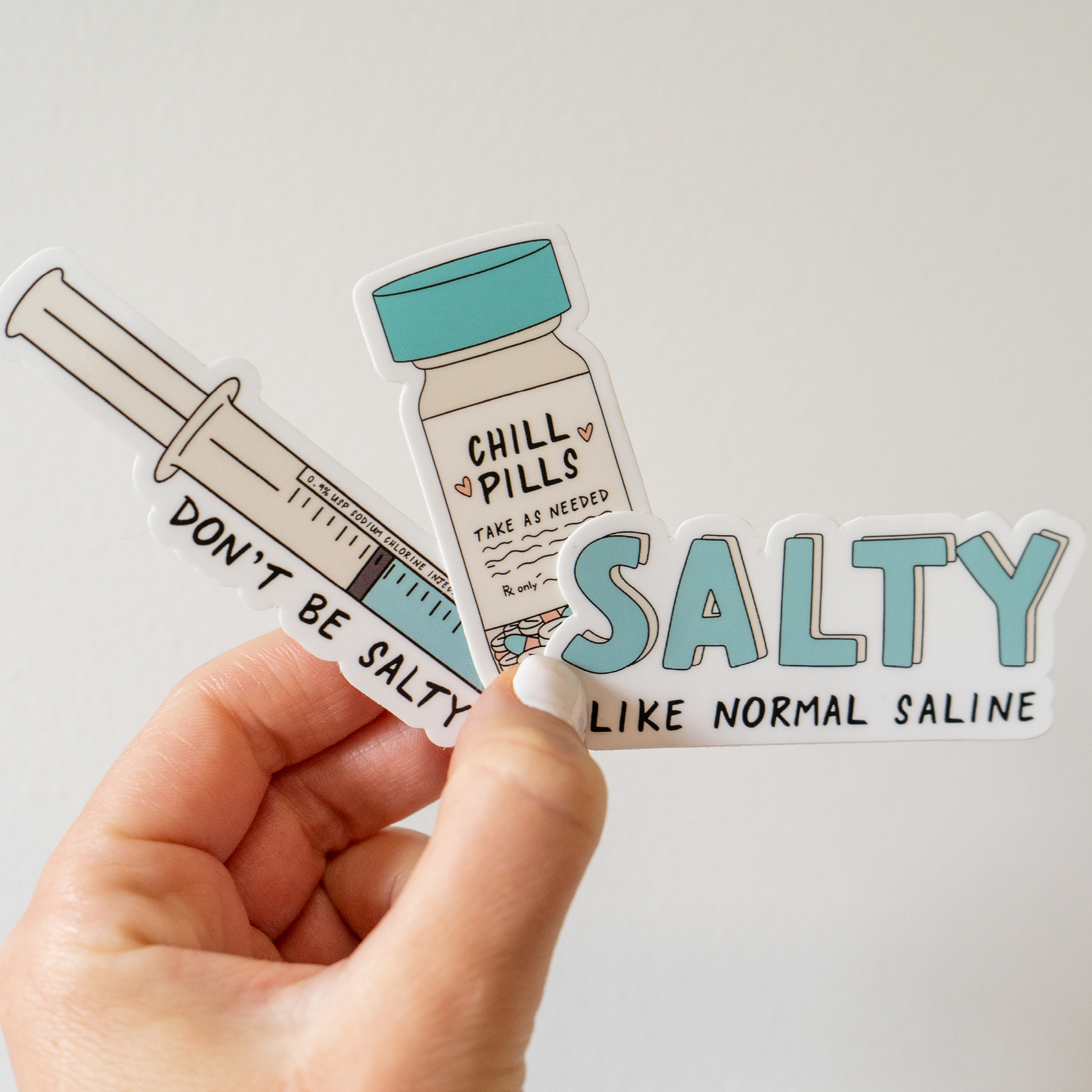 A hand holds a nursing-themed sticker set against a plain background. The stickers feature a syringe labeled “Don’t be salty,” a bottle labeled “Chill pills - take as needed,” and bold text reading “SALTY like normal saline,” referencing nursing humor related to IV fluids.