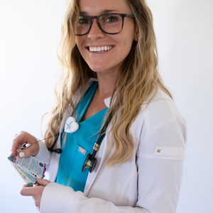 A smiling nurse with long blonde hair, wearing teal scrubs and a white lab coat, holds a set of badge reference cards. A stethoscope with a heart-shaped badge reel is clipped to her coat. She is looking at the camera against a plain white background.