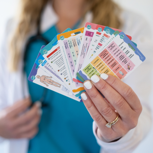 A nurse in a teal scrub top and white coat holds a fanned-out set of colorful badge reference cards. The cards contain essential medical and nursing information. The nurse’s nails are painted white, and a gold ring is visible on one hand.