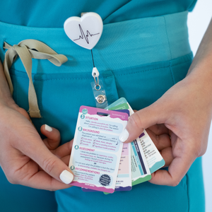 A nurse wearing teal scrubs holds a set of badge reference cards attached to a retractable badge reel with a heart and EKG line design. The cards display medical and nursing information in a compact format. The nurse’s nails are painted white, and a tied drawstring is visible on the scrubs.