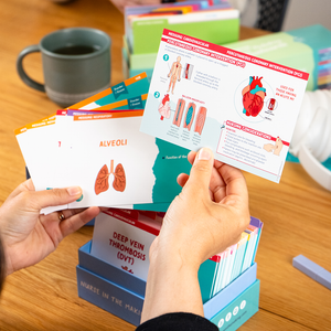 Two people holding Med Surg flashcards at a study table, focusing on a card about “Respiratory Compromise in Post-Op” with anatomical illustrations. Other flashcards, a coffee cup, and study materials are visible in the background.