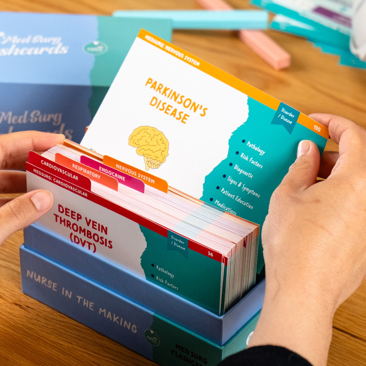 A student holding a Med Surg flashcard about “Parkinson’s Disease” while browsing through a set of organized flashcards in a blue storage box. Another student’s hand is visible assisting, with study materials and other flashcard sets in the background.