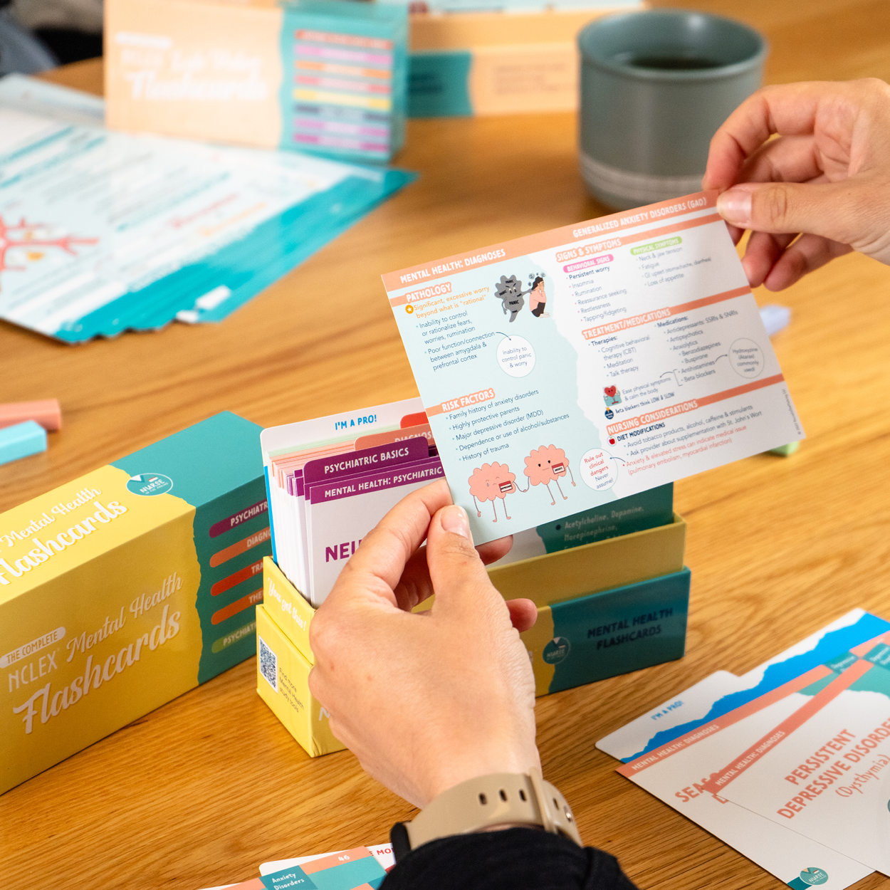 A student studying an NCLEX Mental Health Flashcard from Nurse In The Making, featuring color-coded sections, diagrams, and illustrations on psychiatric concepts. The background includes the full flashcard set, storage box, and study materials on a wooden table.