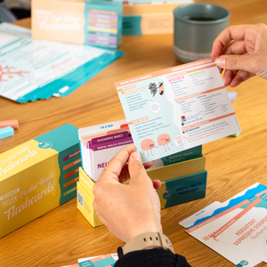 A student studying an NCLEX Mental Health Flashcard from Nurse In The Making, featuring color-coded sections, diagrams, and illustrations on psychiatric concepts. The background includes the full flashcard set, storage box, and study materials on a wooden table.
