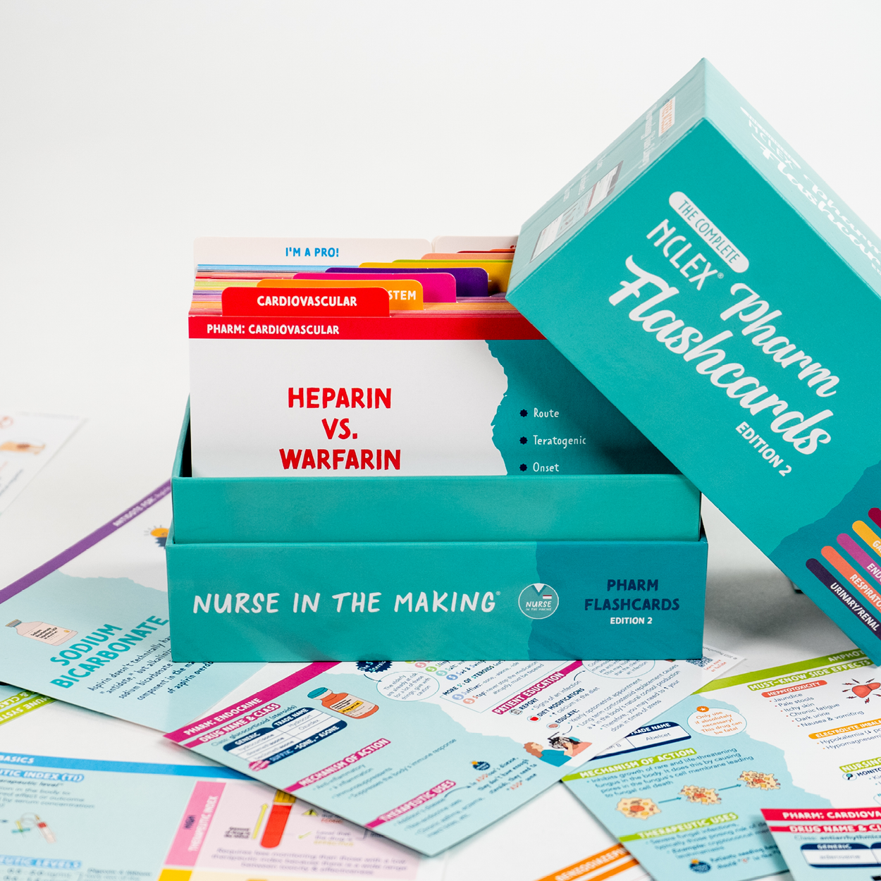 An open box of NCLEX Pharmacology Flashcards, Edition 2, with the lid leaning against the side. The visible flashcard on top is labeled “Heparin vs. Warfarin.” The box is teal and labeled “Nurse in the Making.” Various flashcards with colorful designs and medication information are scattered around the box.