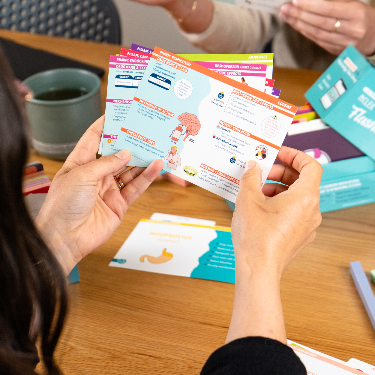 A student holds a set of pharmacology flashcards, focusing on a colorful card with information on a neurological medication. The card includes a brain illustration and key facts highlighted in orange, blue, and white. In the background, another student is holding flashcards, and a teal box labeled “Pharm Flashcards” is open on a wooden table surrounded by study materials.