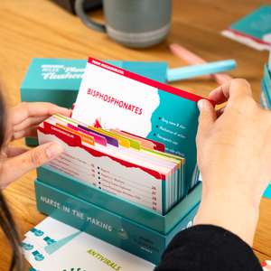 A student flips through a set of pharmacology flashcards, pulling out a card labeled “Bisphosphonates.” The flashcards are neatly organized in a teal box labeled “Pharm Flashcards.” A cup and various study materials, including other flashcards and colored pens, are scattered on a wooden table in the background.