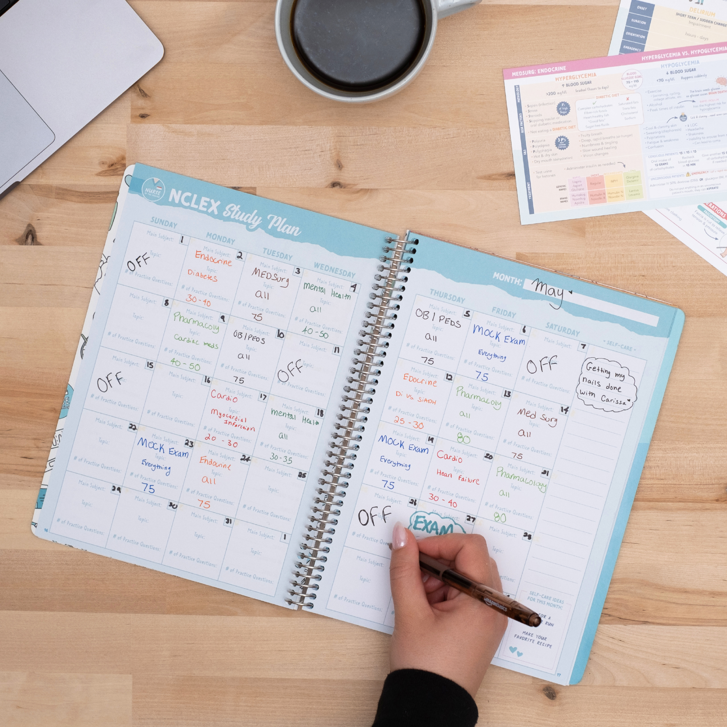 A person writing in an open NCLEX Study Notebook, filling out a structured monthly study plan. The planner features a calendar layout with study topics, notes, and exam preparation schedules. A cup of coffee, a laptop, and additional study materials are on the wooden desk, creating a focused study environment.