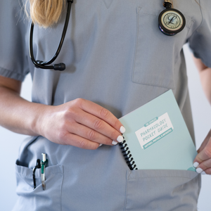 A student wearing gray scrubs and a stethoscope around their neck places the “Pharmacology Pocket Guide” into their front pocket. The spiral-bound guide has a pastel green cover with a white label. Pens are clipped to the scrub pocket. The background is blurred.