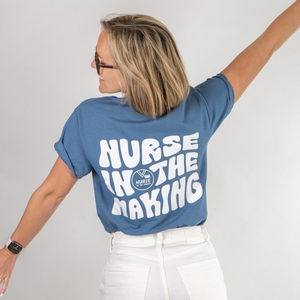 A woman with shoulder-length blonde hair wearing a blue t-shirt with bold white text on the back that reads “Nurse in the Making” along with a small logo. She is playfully stretching her arms out and looking to the side, paired with white pants and a smartwatch. The background is a clean, white studio setting.