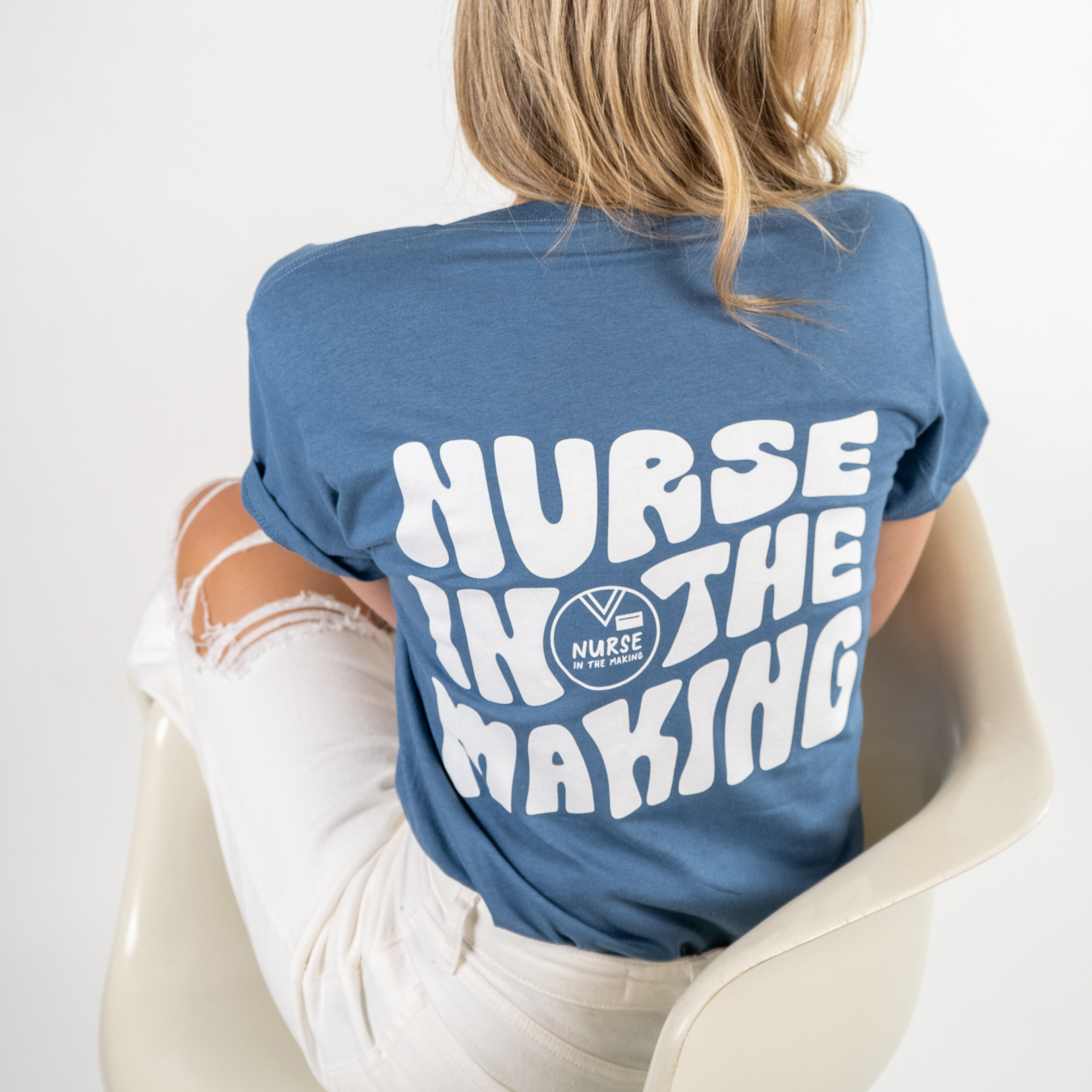 A woman sitting on a modern white chair, wearing a blue t-shirt with bold white text on the back that reads “Nurse in the Making” along with a small logo. She is facing away from the camera, showcasing the design, and is wearing white ripped jeans. The background is a clean, white studio setting.