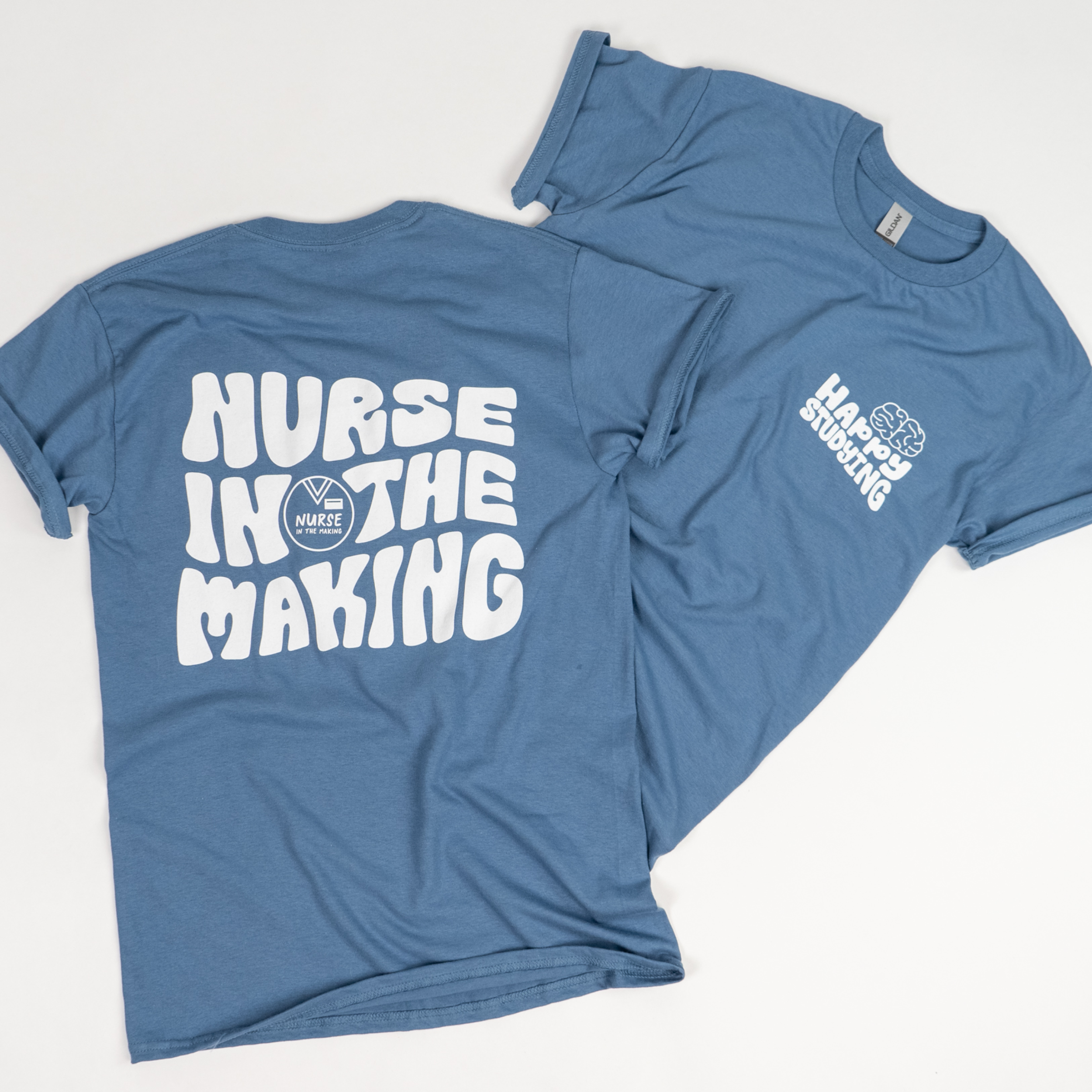 A pair of blue t-shirts laid flat on a white surface. One shirt is displayed with the front side up, featuring a small white logo on the upper left chest that reads “Happy Studying” with a brain icon. The other shirt is flipped to show the back design, which has bold white text reading “Nurse in the Making” along with a small logo.