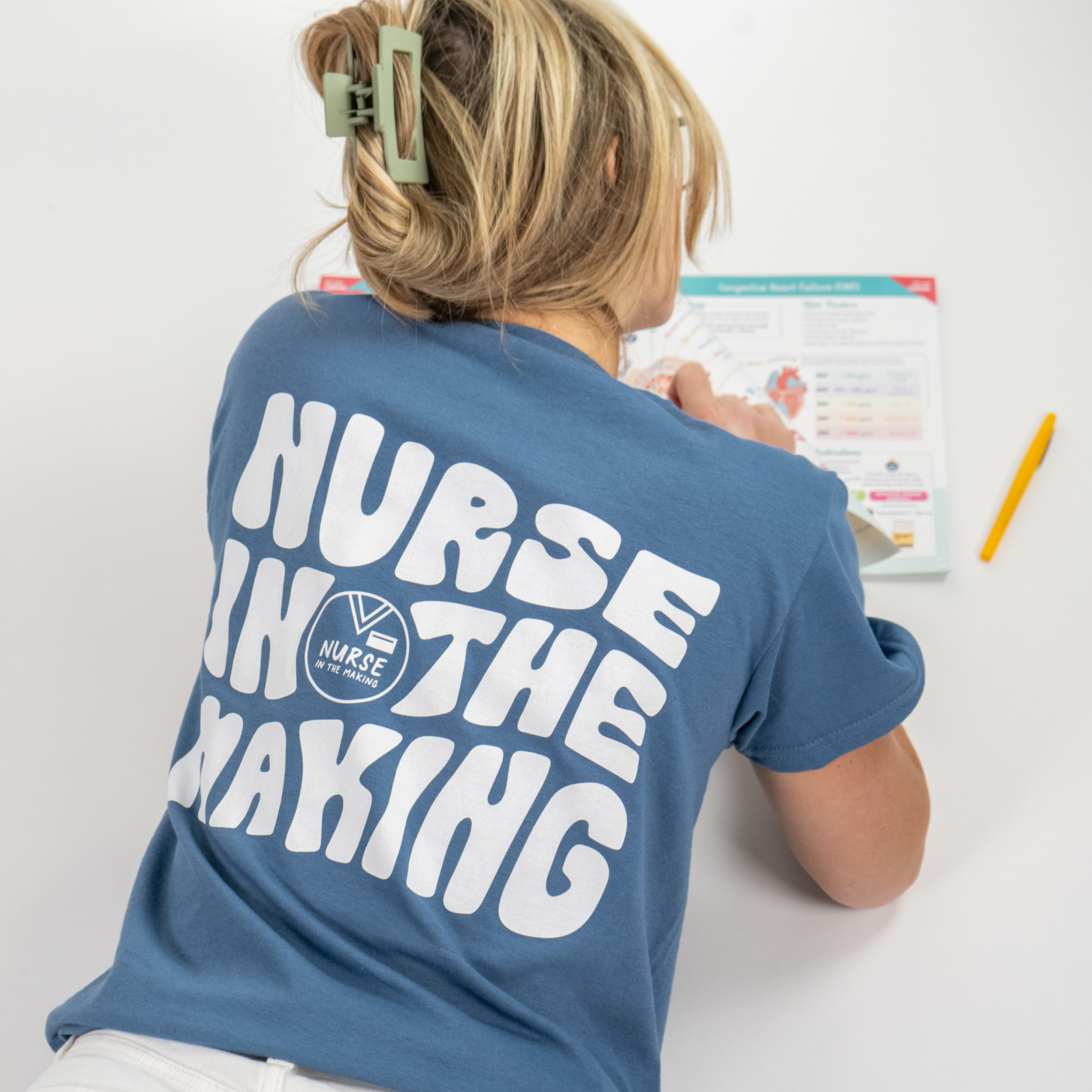 A student wearing a blue t-shirt with bold white text on the back that reads “Nurse in the Making” along with a small logo. She is seated at a desk studying an anatomy guide, with a pencil resting nearby. Her blonde hair is tied up in a casual clip, and she is wearing white pants. The background is a clean, white studio setting.