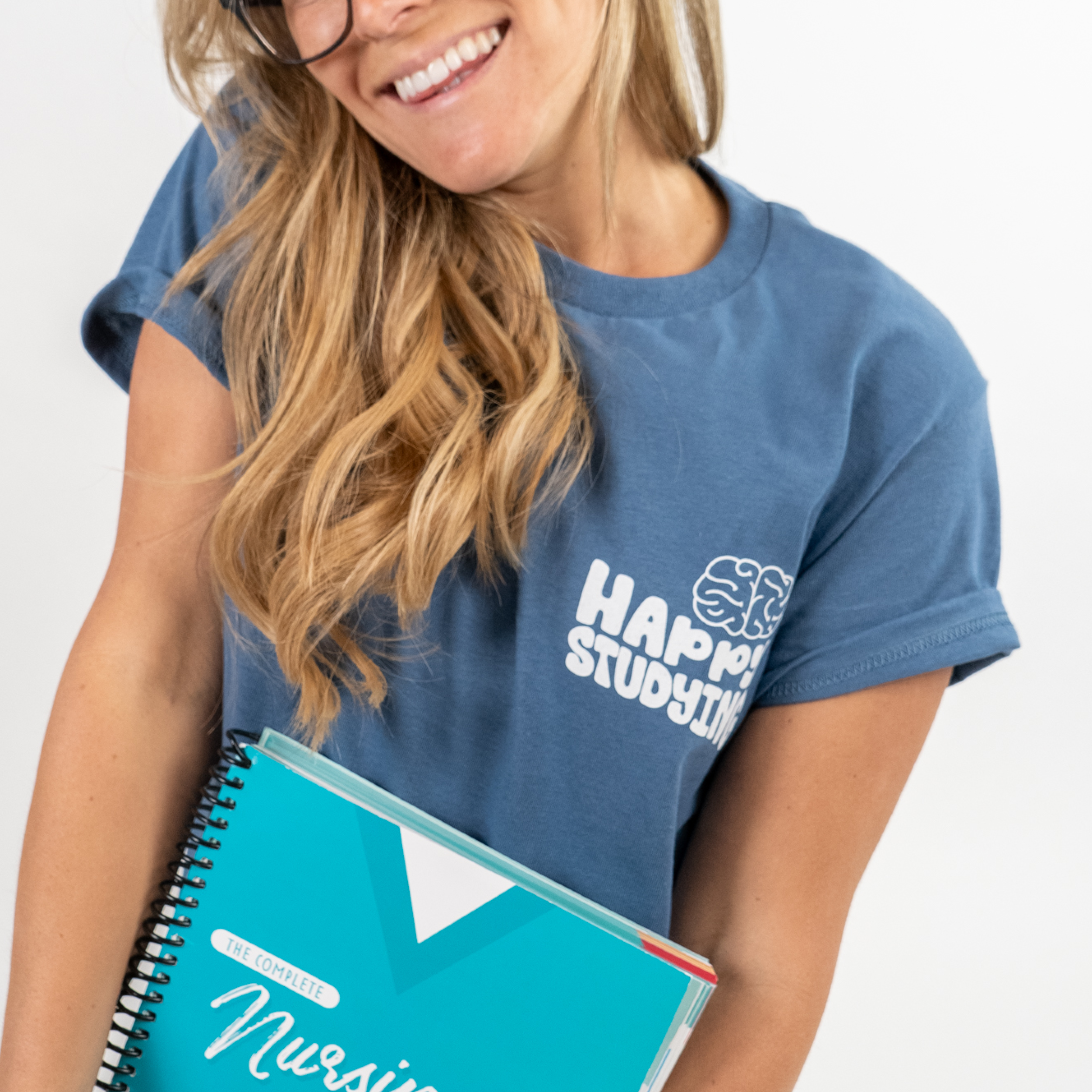A woman with long, wavy blonde hair and glasses smiling while holding The Complete Nursing School Bundle. She is wearing a blue t-shirt with the phrase “Happy Studying” and a brain icon printed on the upper left chest. The background is a clean, white studio setting.