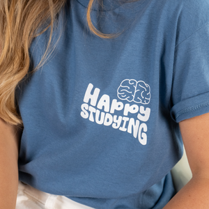 A close-up of a blue t-shirt featuring the phrase “Happy Studying” in bold white text, accompanied by a brain icon. The fabric is soft, with rolled-up sleeves. A portion of long, wavy blonde hair and white pants are visible in the frame. The background is a clean, neutral setting.