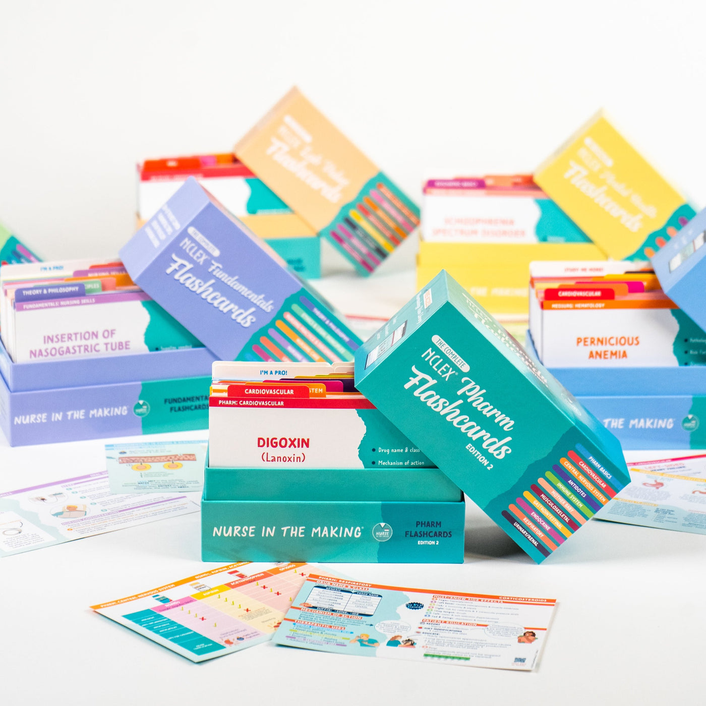 A collection of nursing flashcard boxes arranged in a colorful display. The foreground features an open teal Pharmacology Flashcards box with “Digoxin” visible on one of the cards. Other boxes, including Med Surg, Lab Values, and Pediatrics, are stacked in the background. Various study materials and individual flashcards are scattered around the display. The scene is brightly lit, highlighting the vibrant packaging and detailed study content.