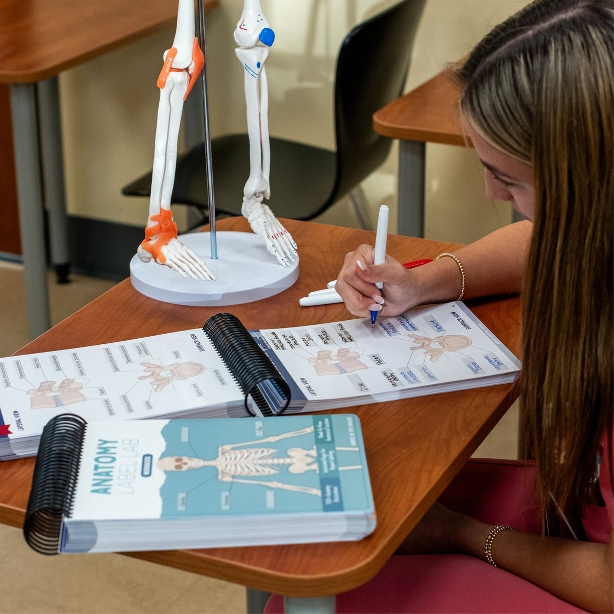 A student in pink scrubs sitting at a classroom desk, using a dry-erase marker to label an anatomy study guide. A spiral-bound anatomy workbook is on the desk, along with a skeletal leg model on a stand. The background features additional desks and chairs in a classroom setting.