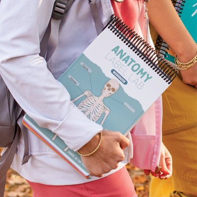 A student is walking while wearing a white jacket, and carrying a backpack holds a spiral-bound Anatomy Label Lab Workbook against their side. The workbook has a teal cover featuring a labeled skeleton diagram. Another student, dressed in colorful scrubs, walks beside them holding additional study materials. The background has a warm, outdoor setting with fallen leaves.