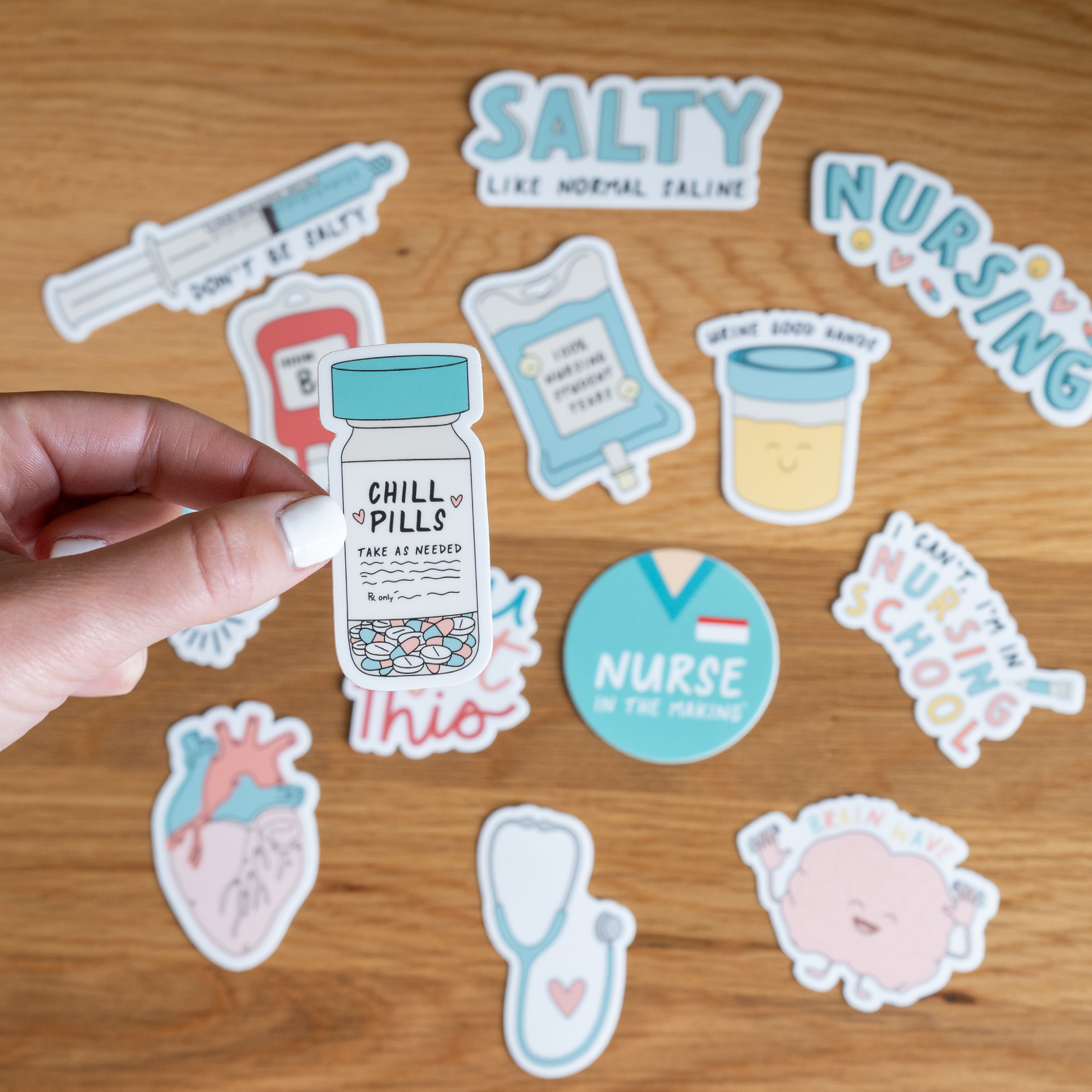 A hand holds a sticker shaped like a pill bottle labeled “Chill pills - take as needed.” In the background, various nursing-themed stickers are spread out on a wooden surface, including designs featuring a heart, brain, IV bag, and humorous medical phrases.