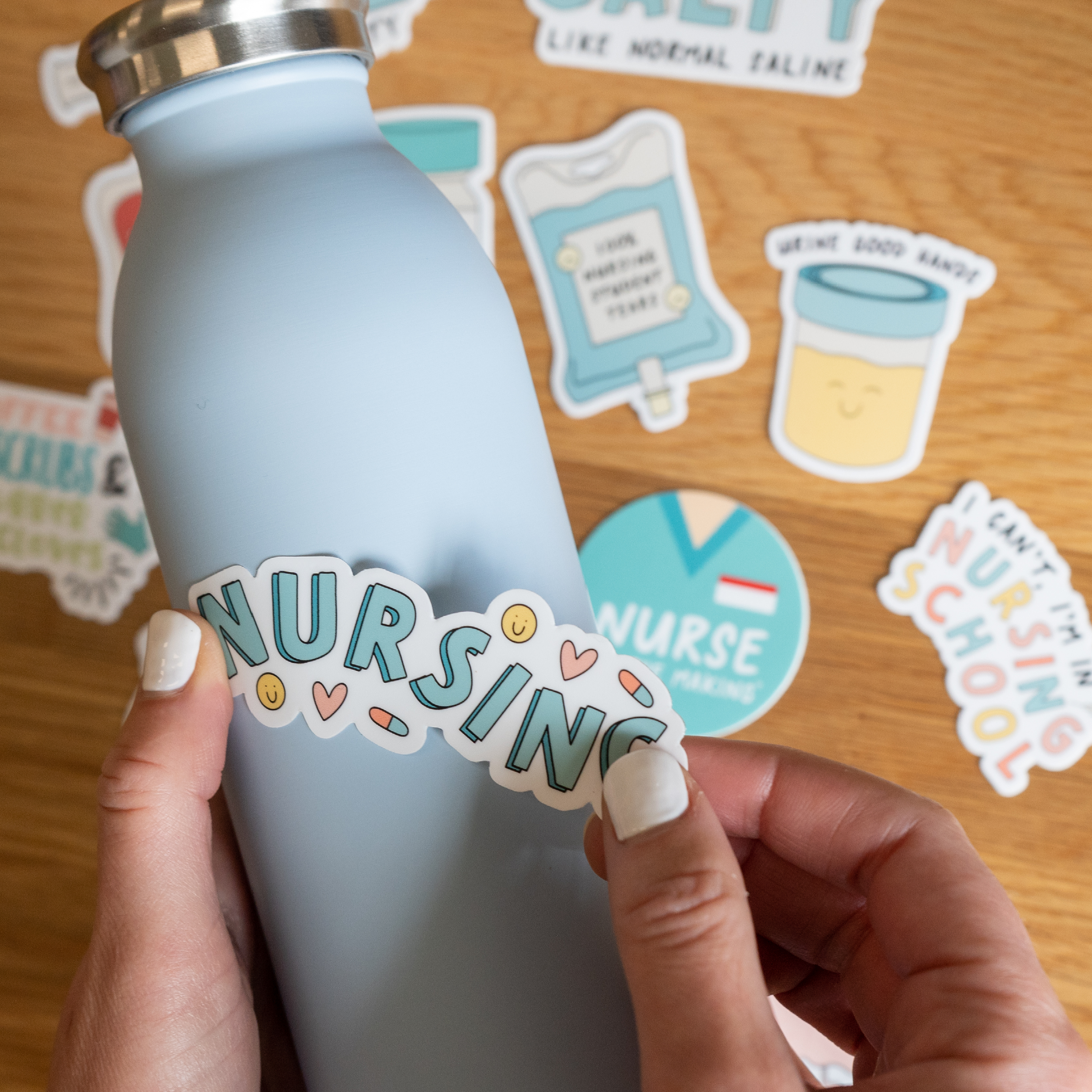 A pair of hands applies a “NURSING” sticker with heart accents to a light blue water bottle. In the background, various nursing-themed stickers, including an IV bag and a “Nurse in the Making” badge, are scattered on a wooden surface.