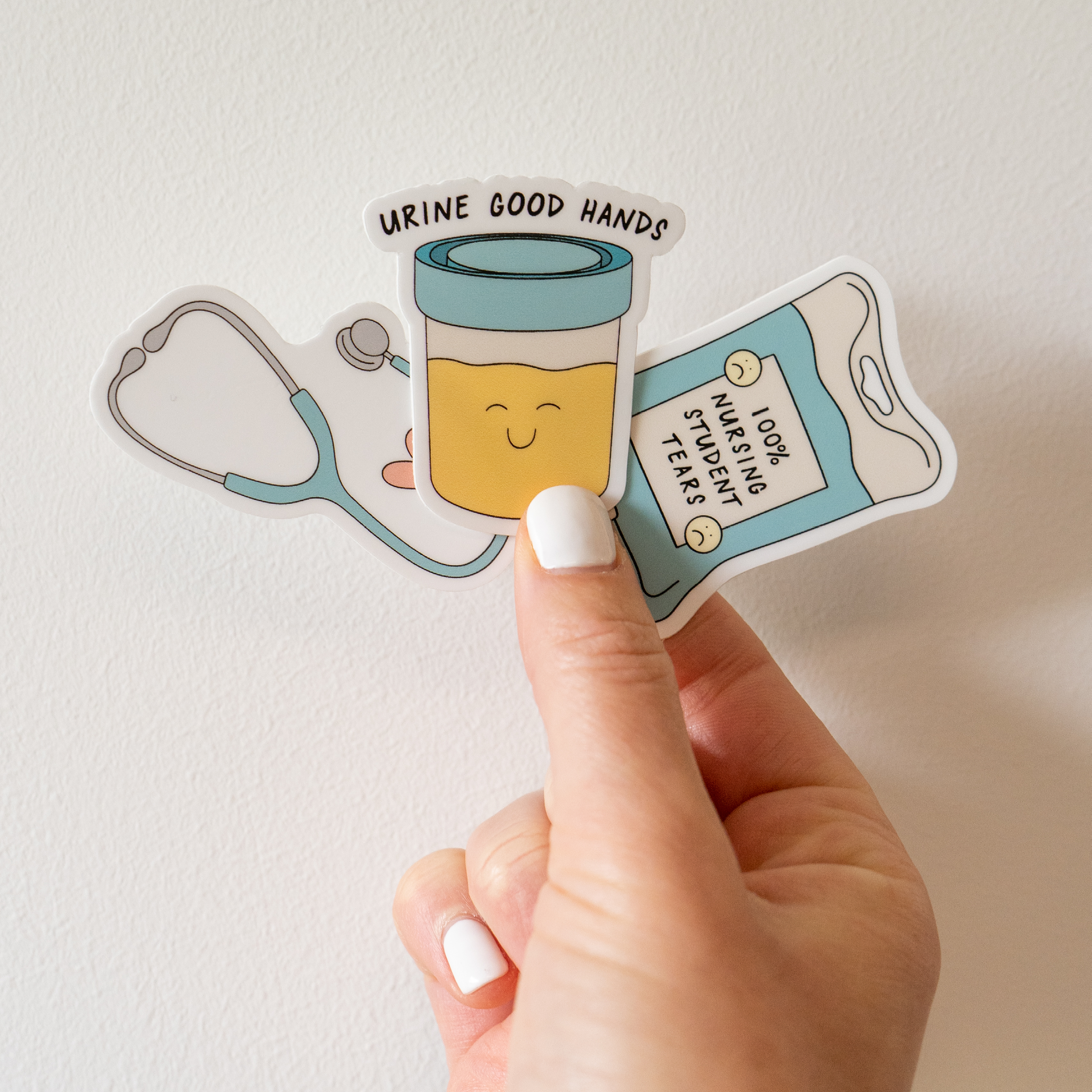 A hand with white-painted nails holds a nursing-themed sticker featuring a smiling urine sample cup with the phrase “Urine good hands,” along with a stethoscope and an IV fluid bag labeled “100% nursing student tears.” The sticker is placed against a plain background.