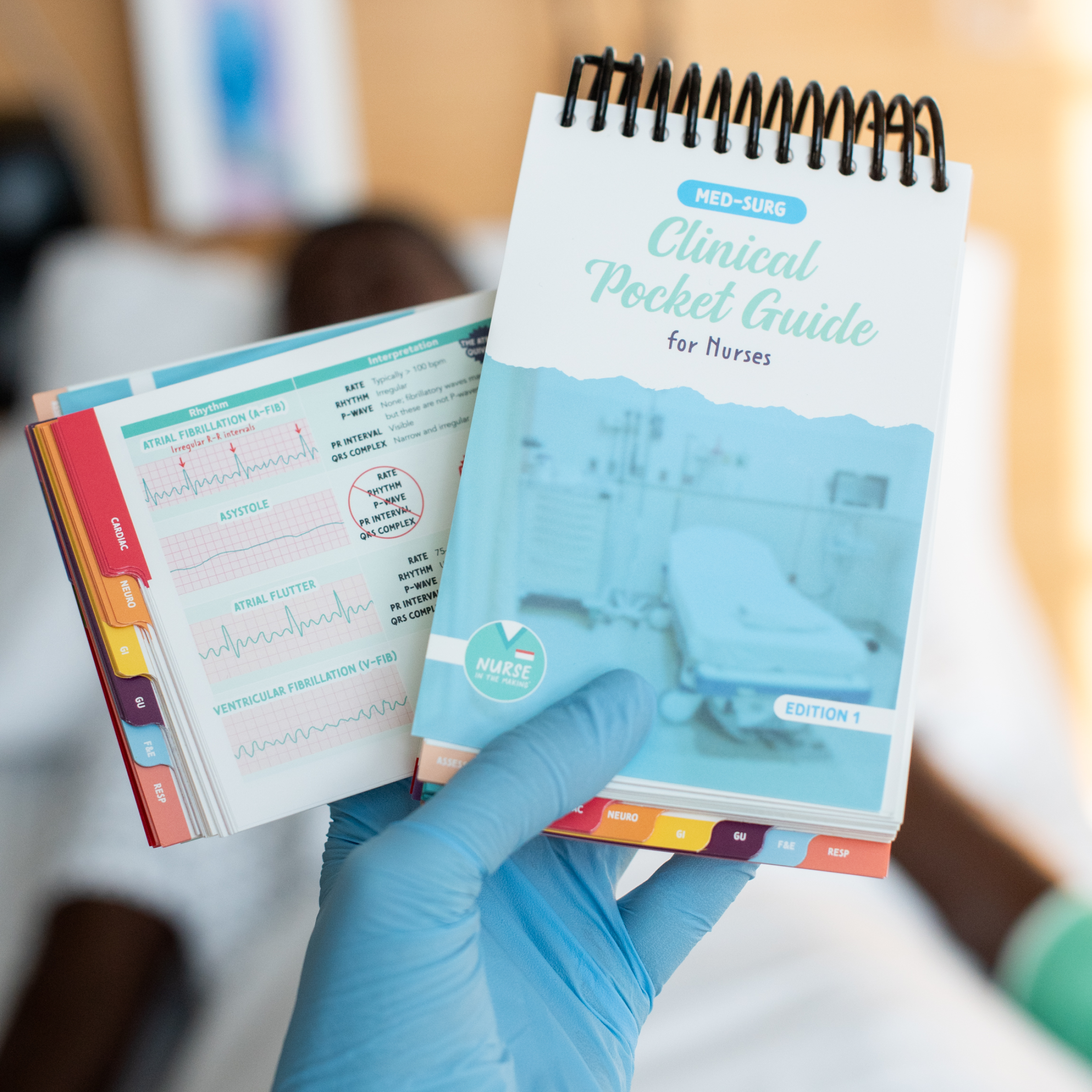 A gloved hand holding a Med-Surg Clinical Pocket Guide for Nurses, a spiral-bound reference book with a blue-toned hospital scene on the cover. The second hand is flipping through an open page displaying colorful ECG rhythm strips and medical notes. The blurred background features a clinical setting with a simulated patient lying in bed.
