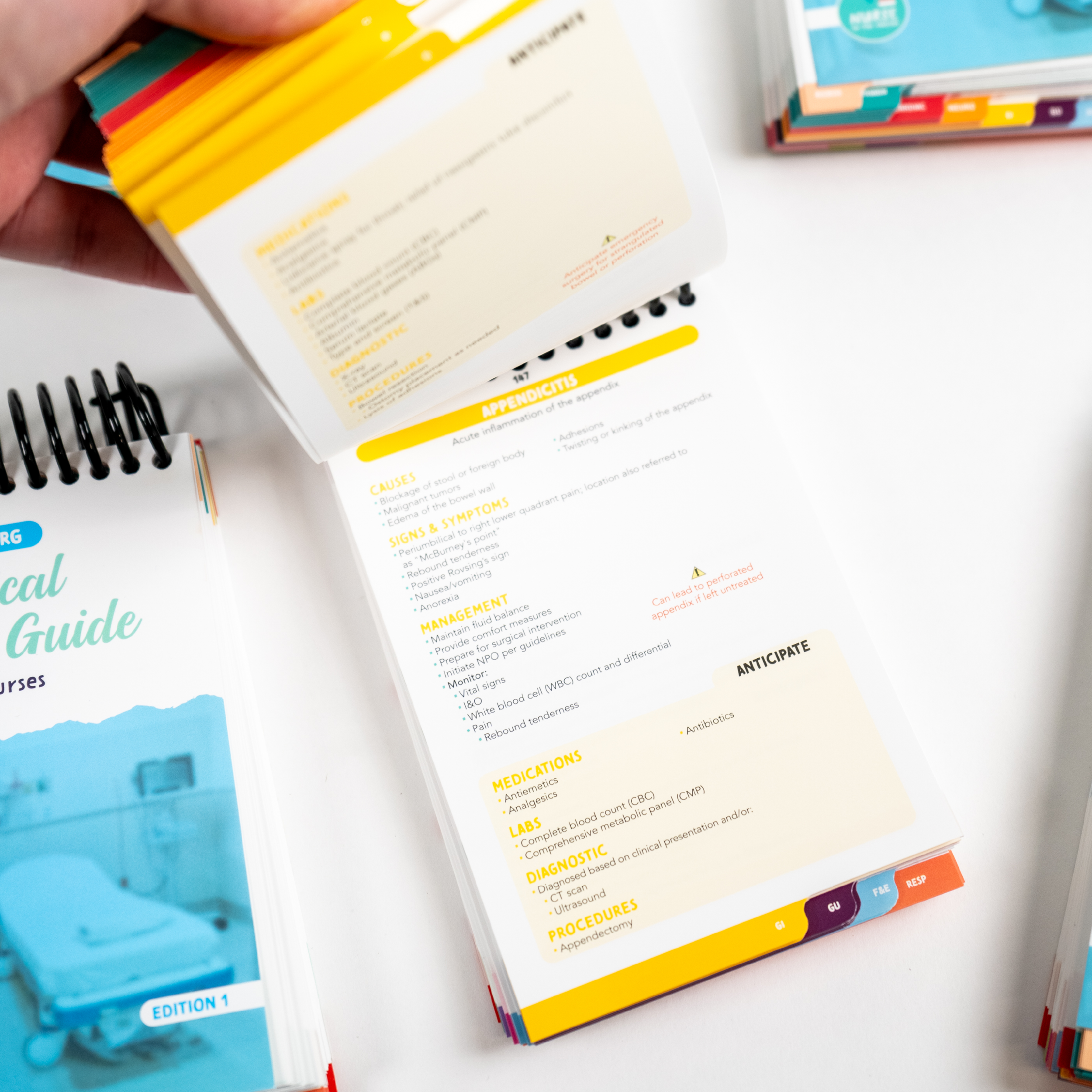 A close-up of a Clinical Pocket Guide for Nurses, with a hand flipping through its spiral-bound pages. The open page features a yellow-highlighted medication reference section with detailed information on indications, side effects, and dosage. Color-coded tabs are visible for easy navigation. Another copy of the guide and its cover are partially visible in the background on a white surface.