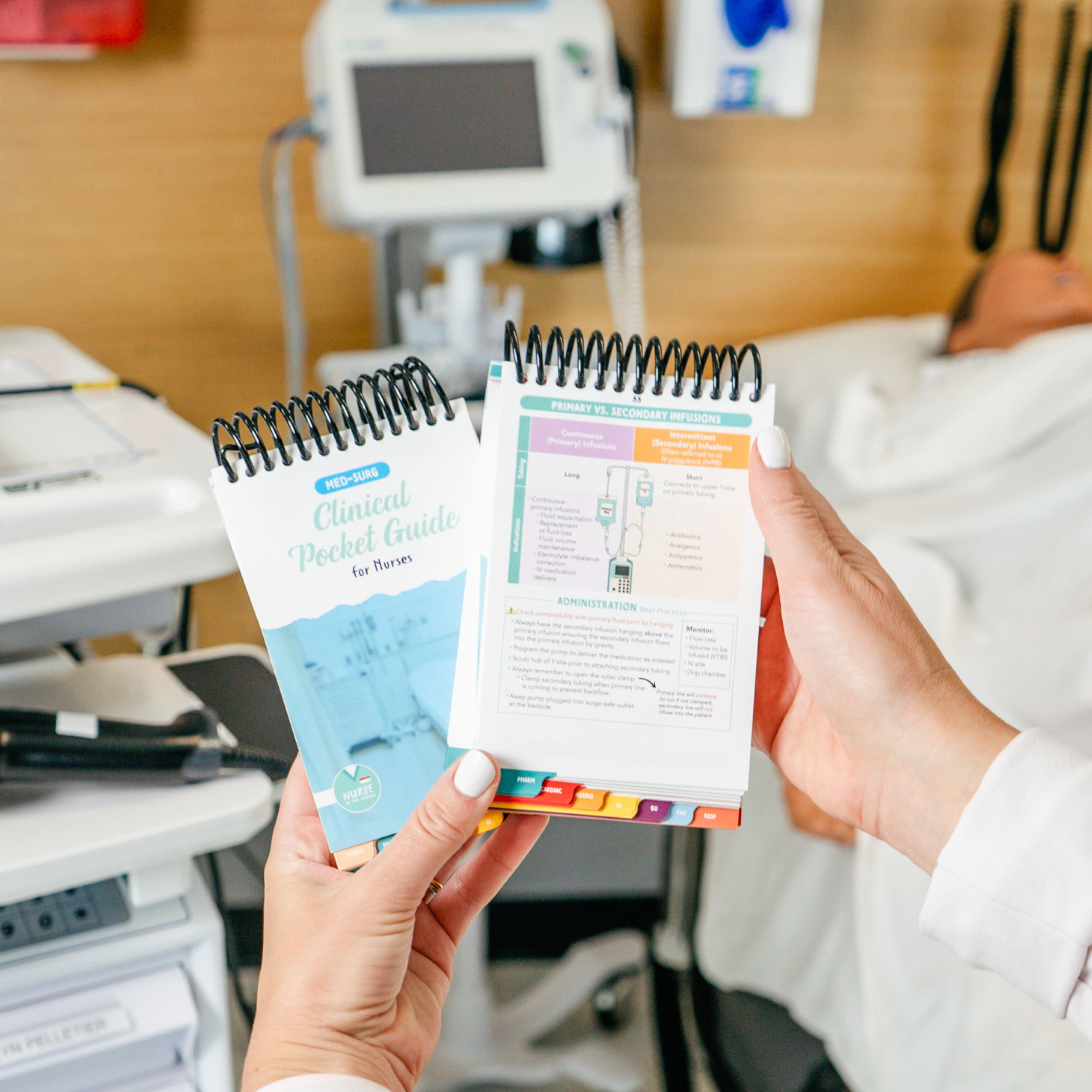 A nurse in a white lab coat holds two Clinical Pocket Guides for Nurses in a hospital setting. One guide is closed, displaying its blue cover with an image of a hospital bed, while the other is open, revealing a reference page with colorful text and diagrams. In the background, there is medical equipment, including a monitor, a medical cart, a simulated patient, and a hospital bed.