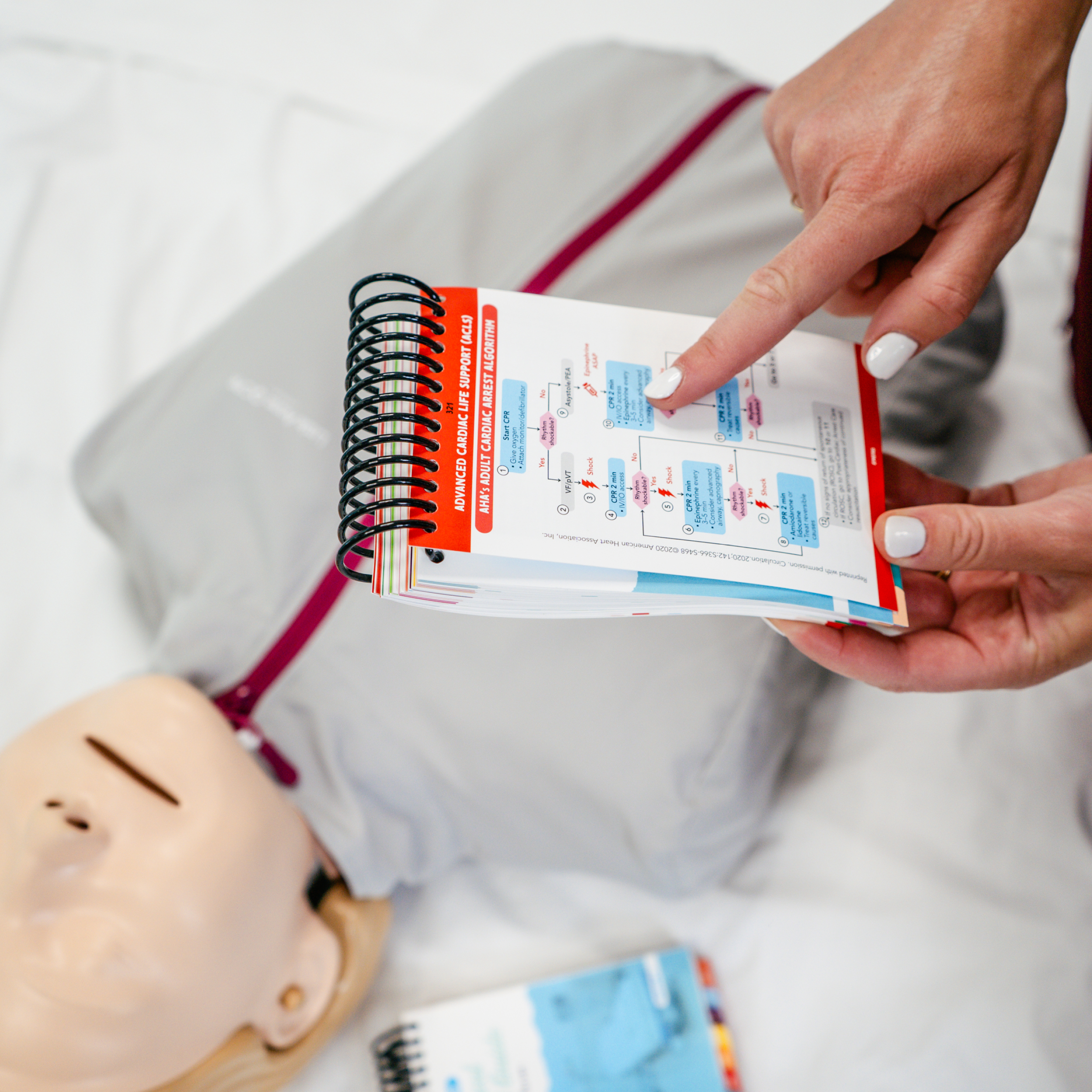 A nurse holds an open Clinical Pocket Guide for Nurses, pointing to a reference page with a red header, flowcharts, and medical information. In the background, a CPR mannequin is lying on a hospital bed, along with another closed pocket guide featuring a blue cover.
