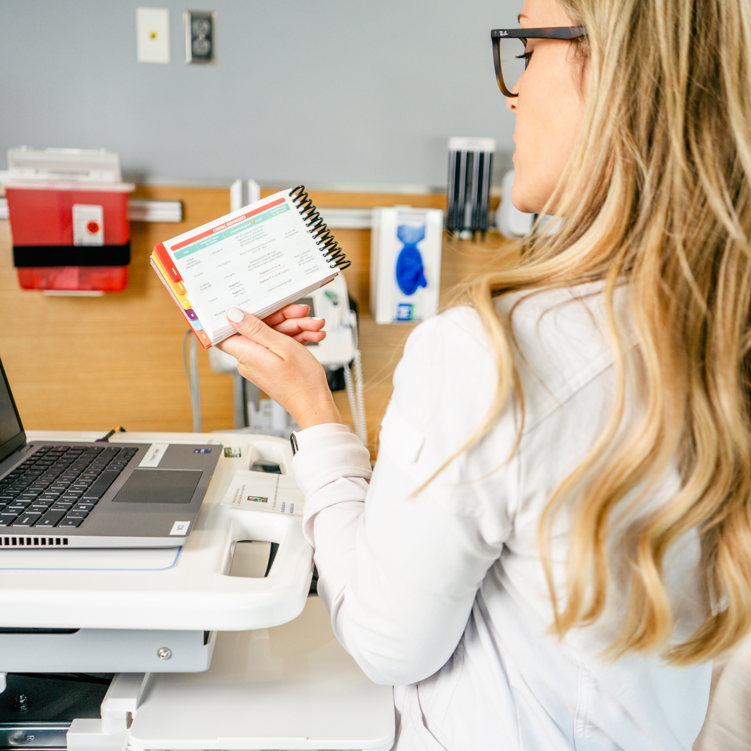 A nurse with long blonde hair, wearing glasses and a white lab coat, is standing at a mobile workstation in a clinical setting. She holds a Clinical Pocket Guide with a spiral binding, open to a reference page with colorful tabs visible on the side. In front of her is a laptop, and the background features medical equipment, gloves, and a red medical waste bin all attached to the wall.