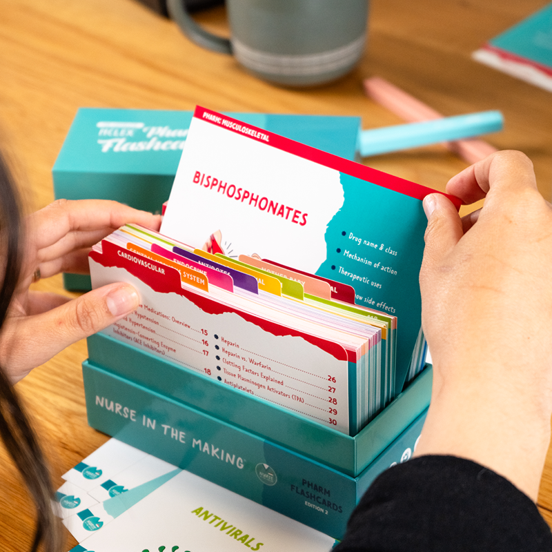 A student holding a set of Pharmacology Flashcards, pulling out a card titled “Bisphosphonates.” The flashcards are neatly organized in a teal storage box labeled “Nurse in the Making.” In the background, a wooden table holds study materials, including additional flashcards, a cup, and colorful pens. The scene is well-lit, emphasizing the detailed and structured content of the flashcards.