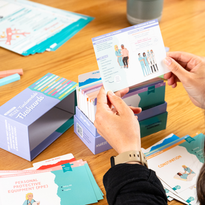 A person holding a NCLEX Fundamentals Flashcard displaying a healthcare-related illustration and key nursing concepts. The background shows an open box of flashcards, study materials, and a wooden desk setup with additional nursing resources.