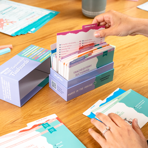 A person going through a box of NCLEX Fundamentals Flashcards on a wooden desk, with study materials, nursing flashcards, and a blue box labeled “Fundamentals Flashcards” in the background.