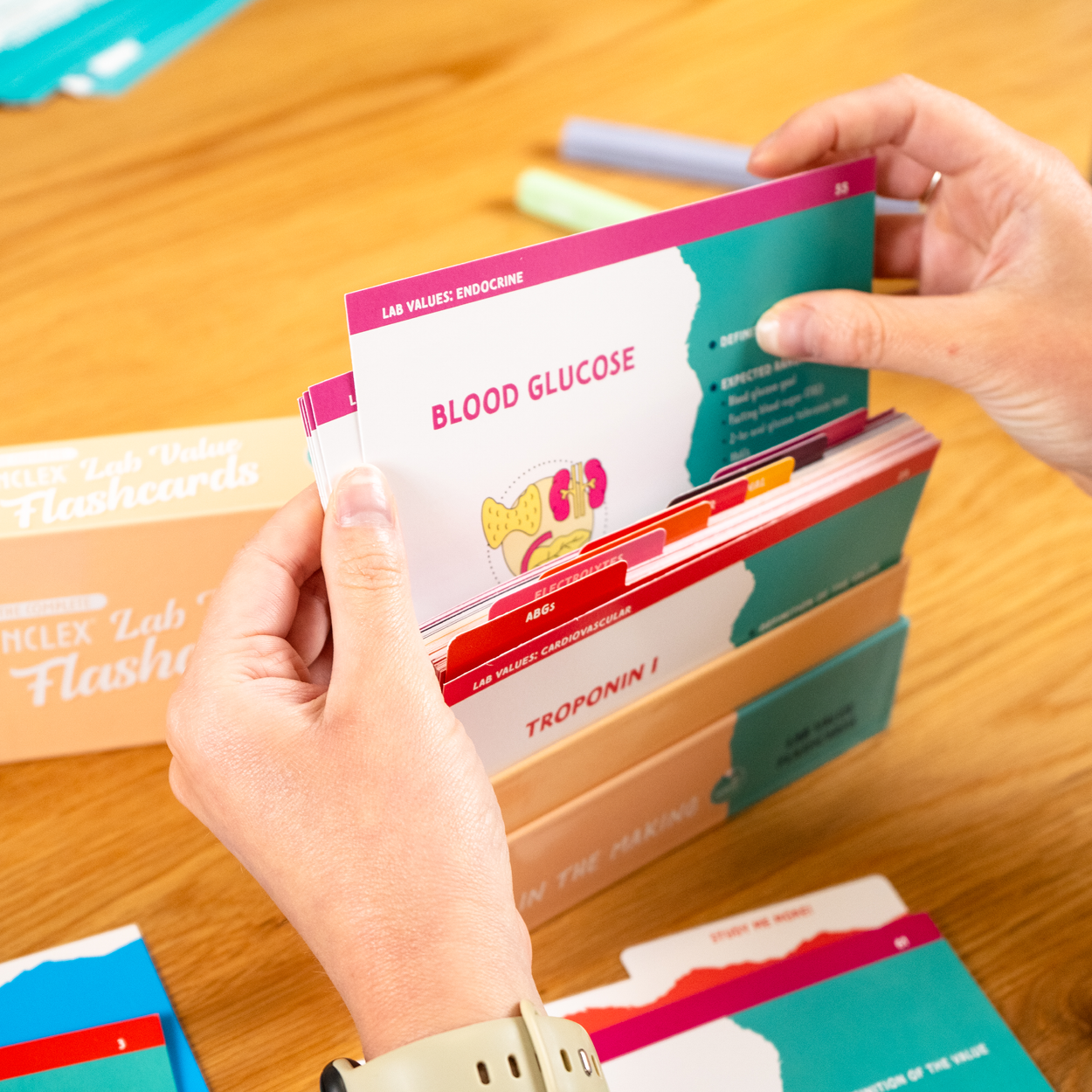 A person flips through a set of Nurse In The Making's NCLEX Lab Value Flashcards, holding a card titled “Blood Glucose.” The card features an illustration and key information about blood glucose levels. The background includes a flashcard storage box labeled “Lab Value Flashcards,” additional study materials, and a neat study setup.