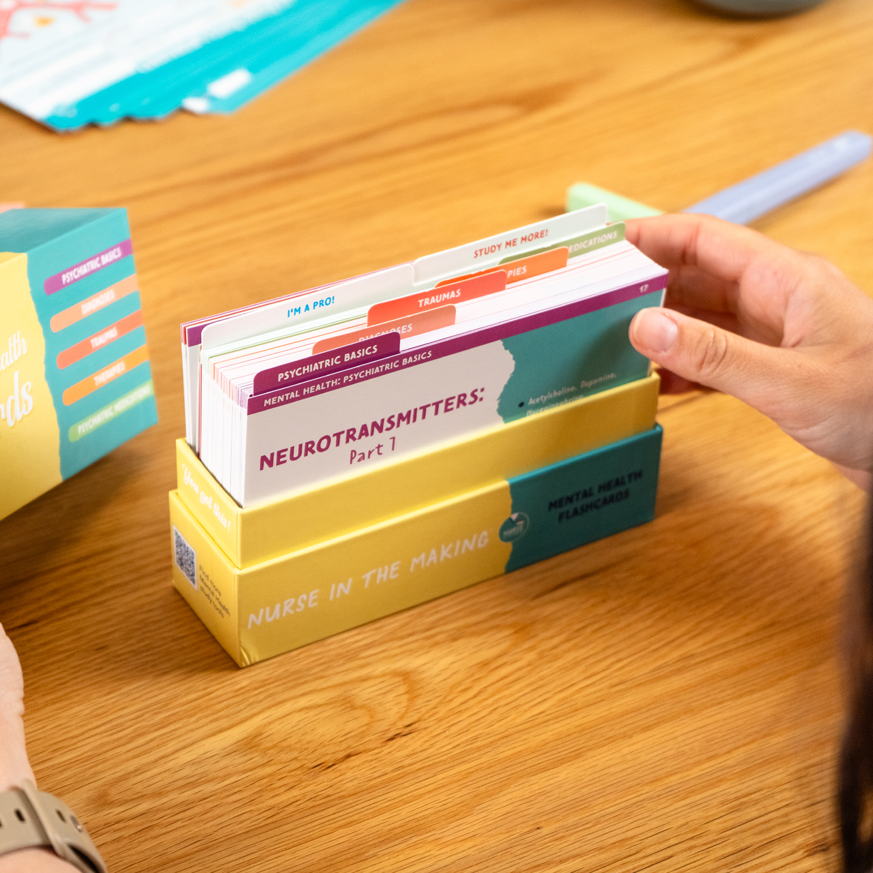 A student's hand holding through the NCLEX Mental Health Flashcards from Nurse in the Making, neatly stored in a pastel yellow branded box. The color-coded sections focus on neurotransmitters and psychiatric basics. A wooden table with study materials is in the background.