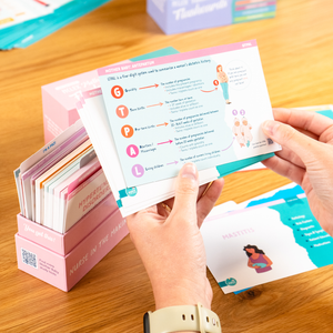 A person holding an NCLEX Mother Baby Flashcard from a pink flashcard set, displaying key prenatal assessment points. The flashcard box and additional study materials are visible on a wooden table in the background.