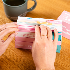 A student flipping through a set of NCLEX Mother Baby Flashcards in a pink box, organized with labeled dividers for different maternity and newborn care topics. A coffee cup and additional flashcard boxes are in the background on a wooden table.