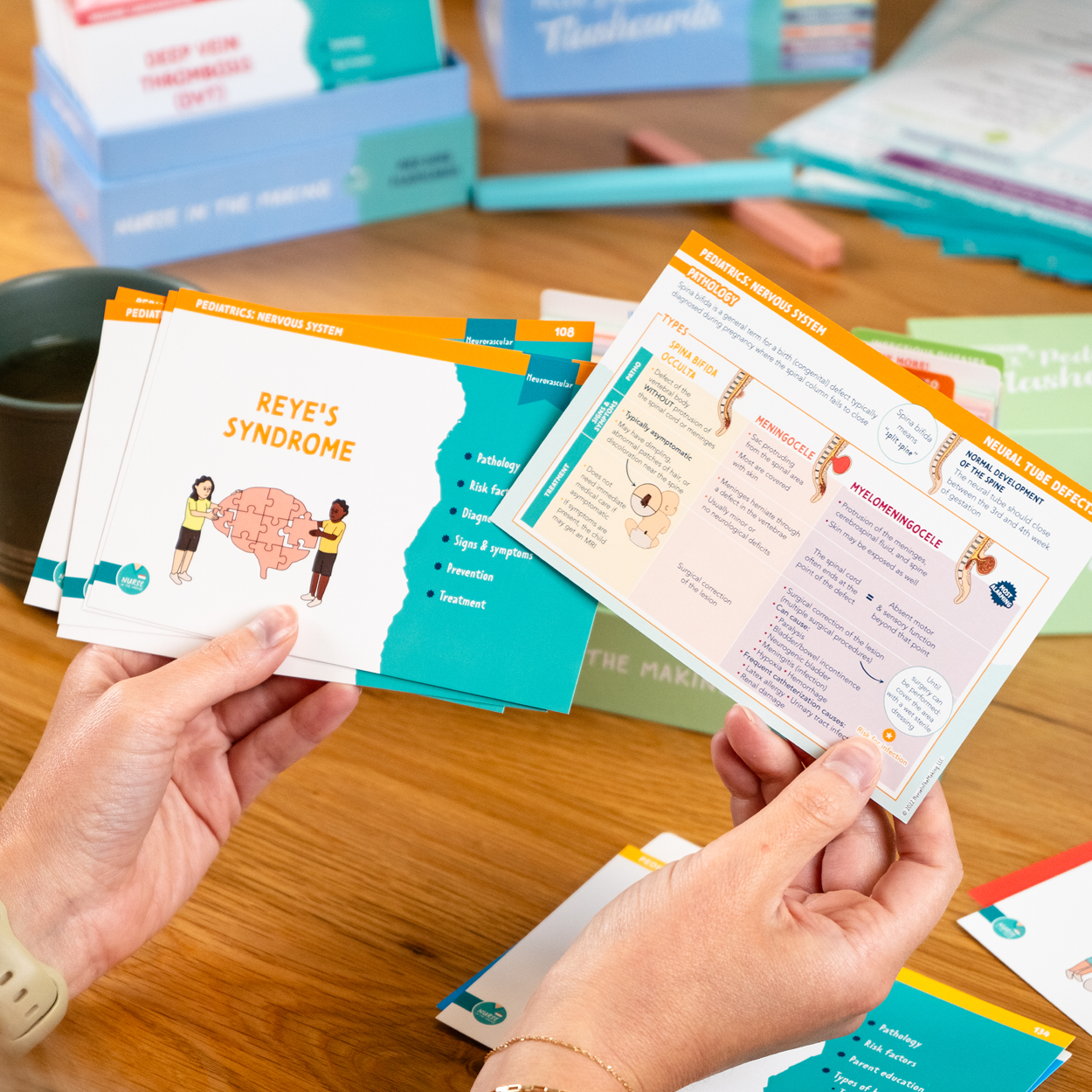 A person holding NCLEX Pediatrics Flashcards focused on Reye’s Syndrome and Meningitis, featuring colorful medical illustrations and key nursing considerations. The background includes an open study setup with additional flashcards, study materials, and storage boxes from Nurse In The Making.