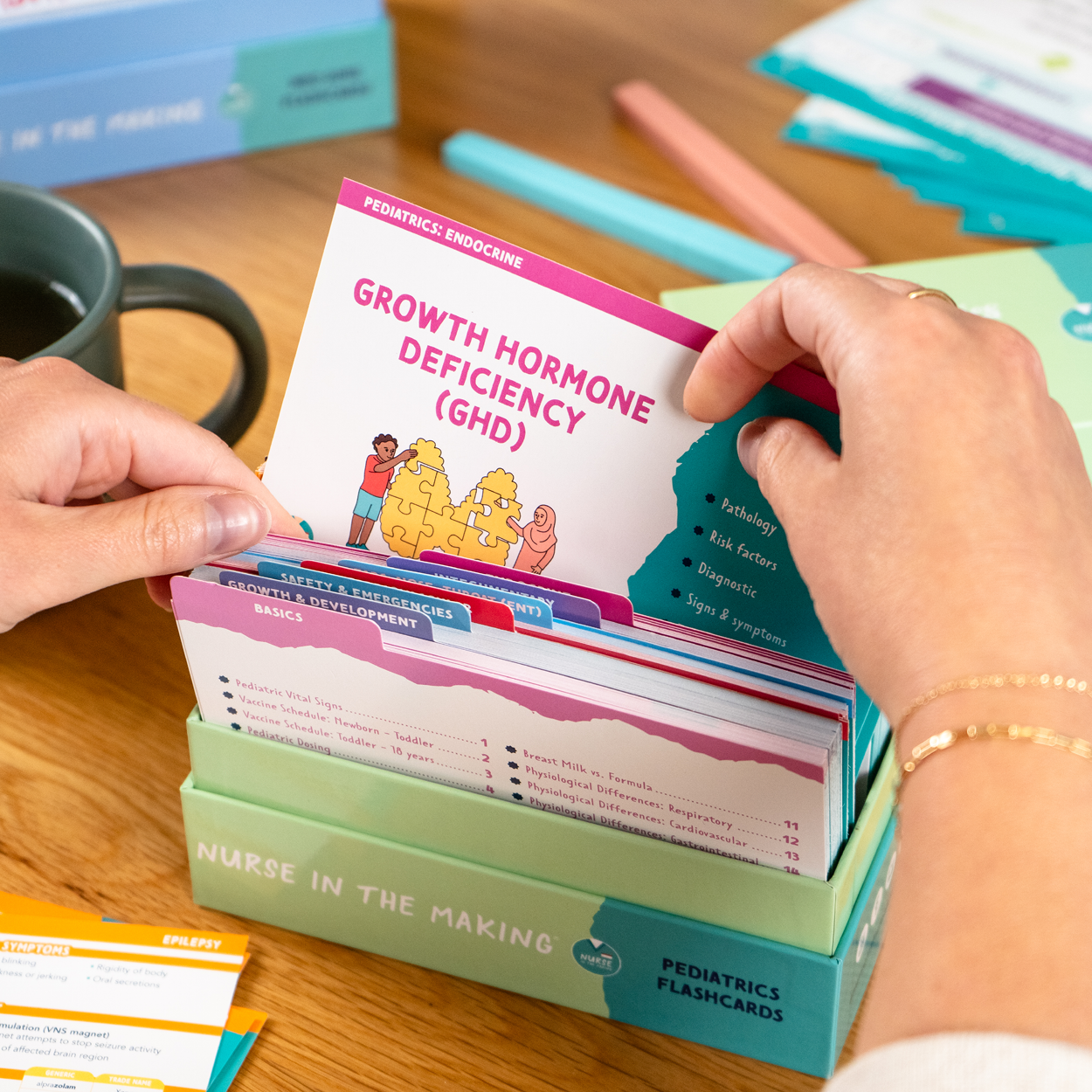 A person flipping through NCLEX Pediatrics Flashcards, highlighting Growth Hormone Deficiency (GHD) with engaging medical illustrations and key information. The background includes a study setup with more flashcards, highlighters and a coffee mug.