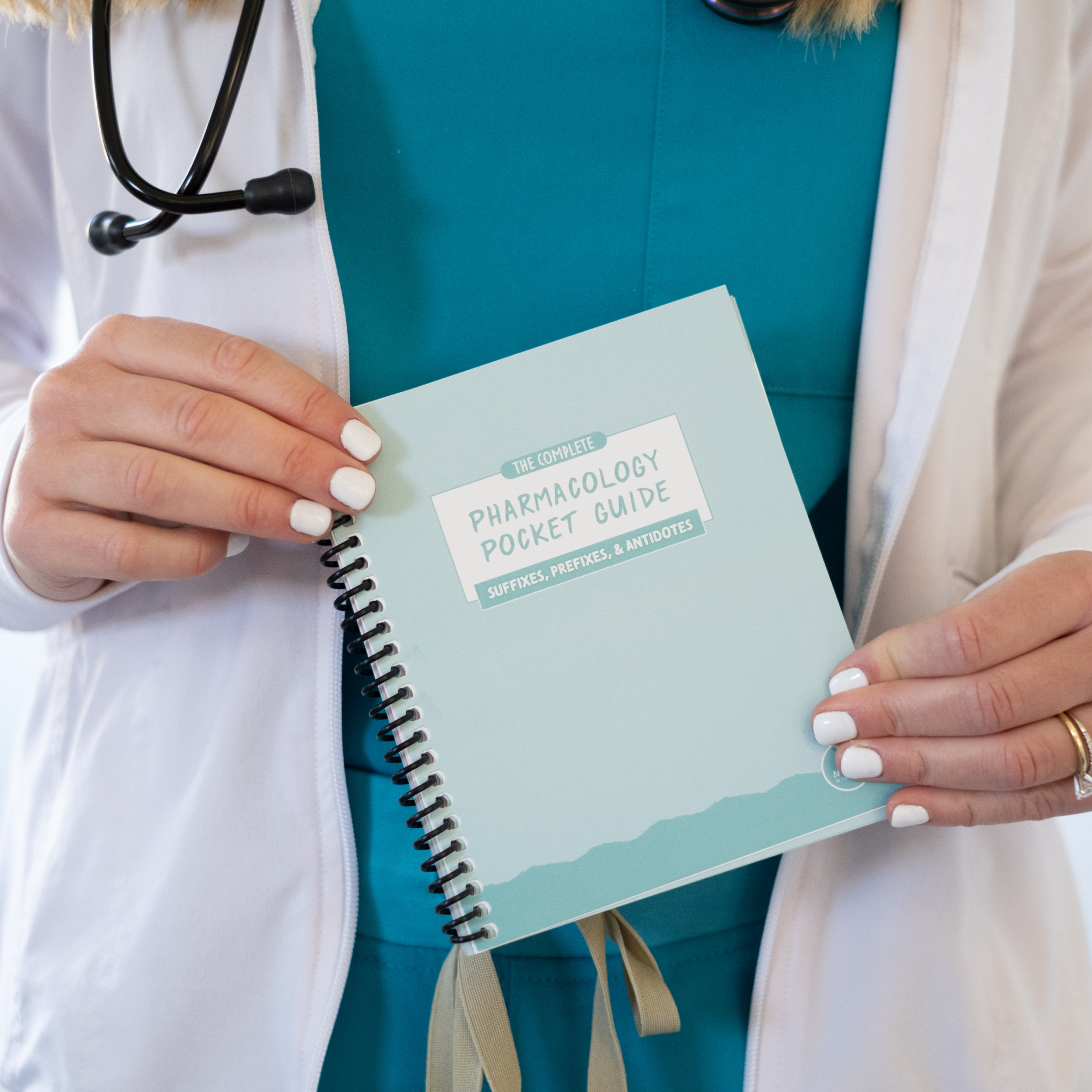 A student wearing teal scrubs and a white lab coat holds the “Pharmacology Pocket Guide” with both hands. A stethoscope hangs around their neck. The spiral-bound guide has a pastel green cover with a white label. The background is blurred.