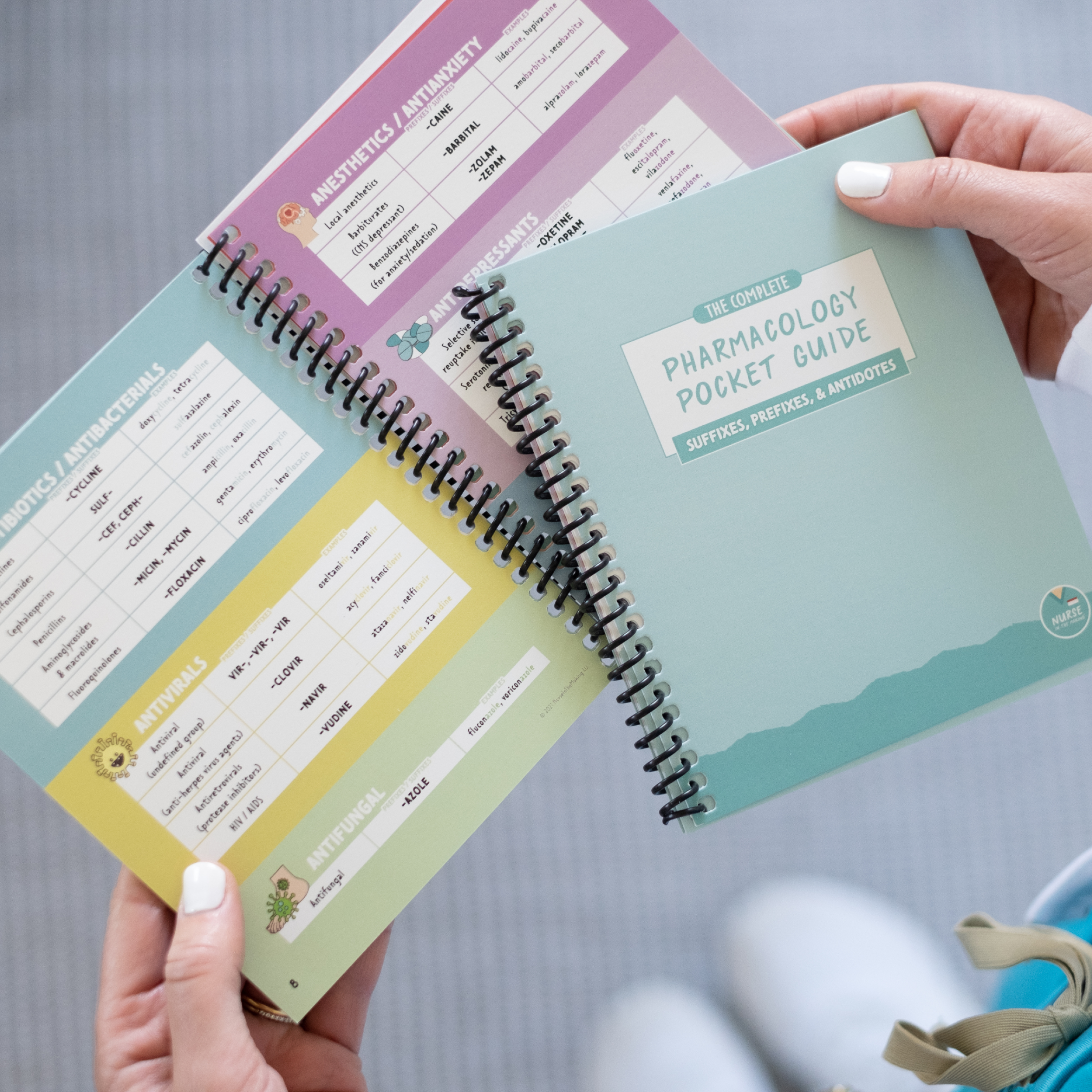 A student holds an open “Pharmacology Pocket Guide” with a pastel green cover in one hand and flips through the spiral-bound pages with the other. The guide displays colorful drug classification charts with dosage details. The background shows the student’s white shoes and a teal backpack strap.