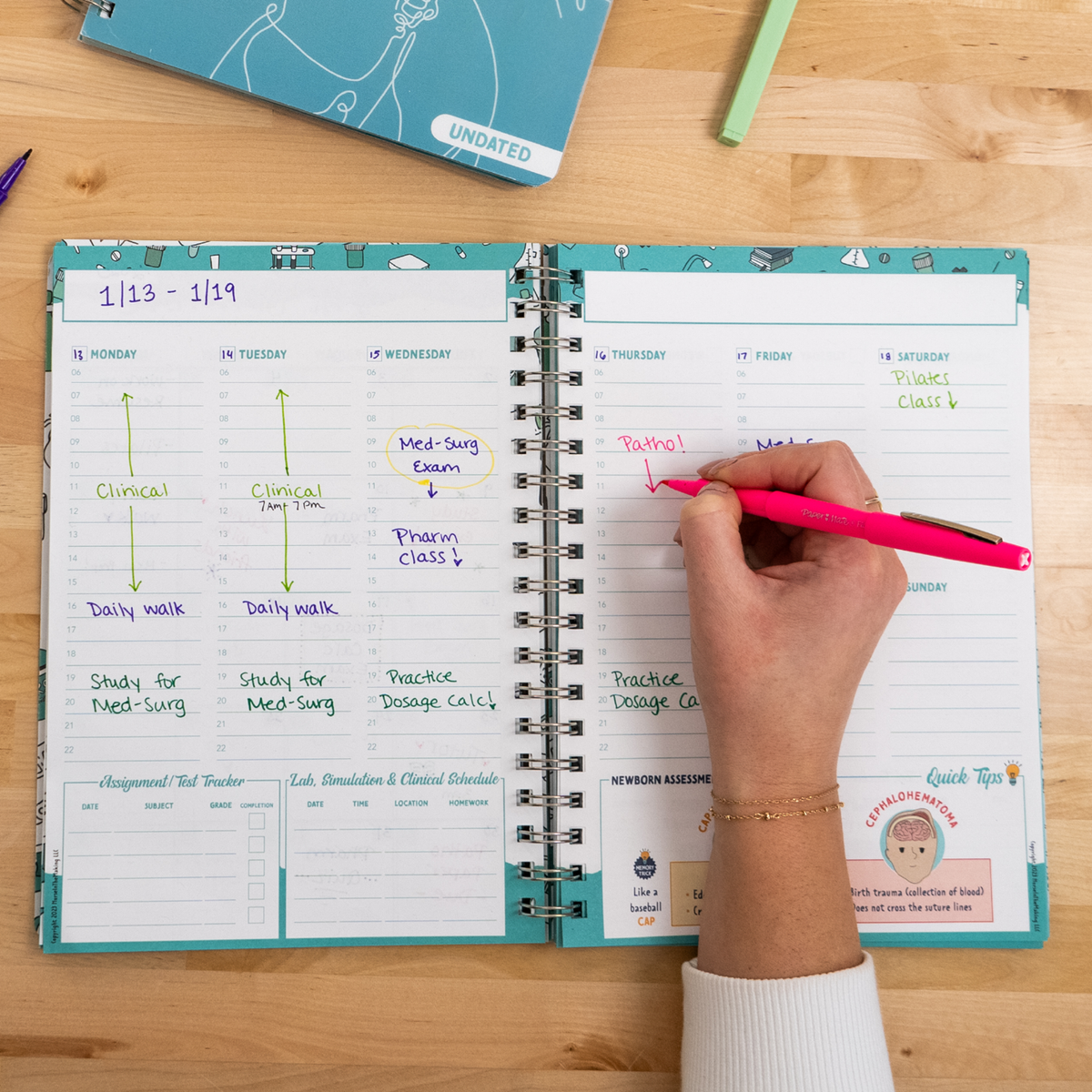 A student writes with a pink pen in a spiral-bound planner open to a weekly schedule. The planner has handwritten notes in various colors detailing study sessions, exams, and assignments. A closed planner and highlighter rest on the wooden desk.