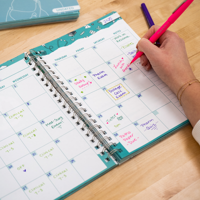 A student writes in a nursing school planner with a pink pen. The open planner displays a monthly calendar filled with handwritten notes in various colors. A teal hardcover planner with a medical-themed design is partially visible in the background on a wooden desk, alongside study materials and a purple pen.