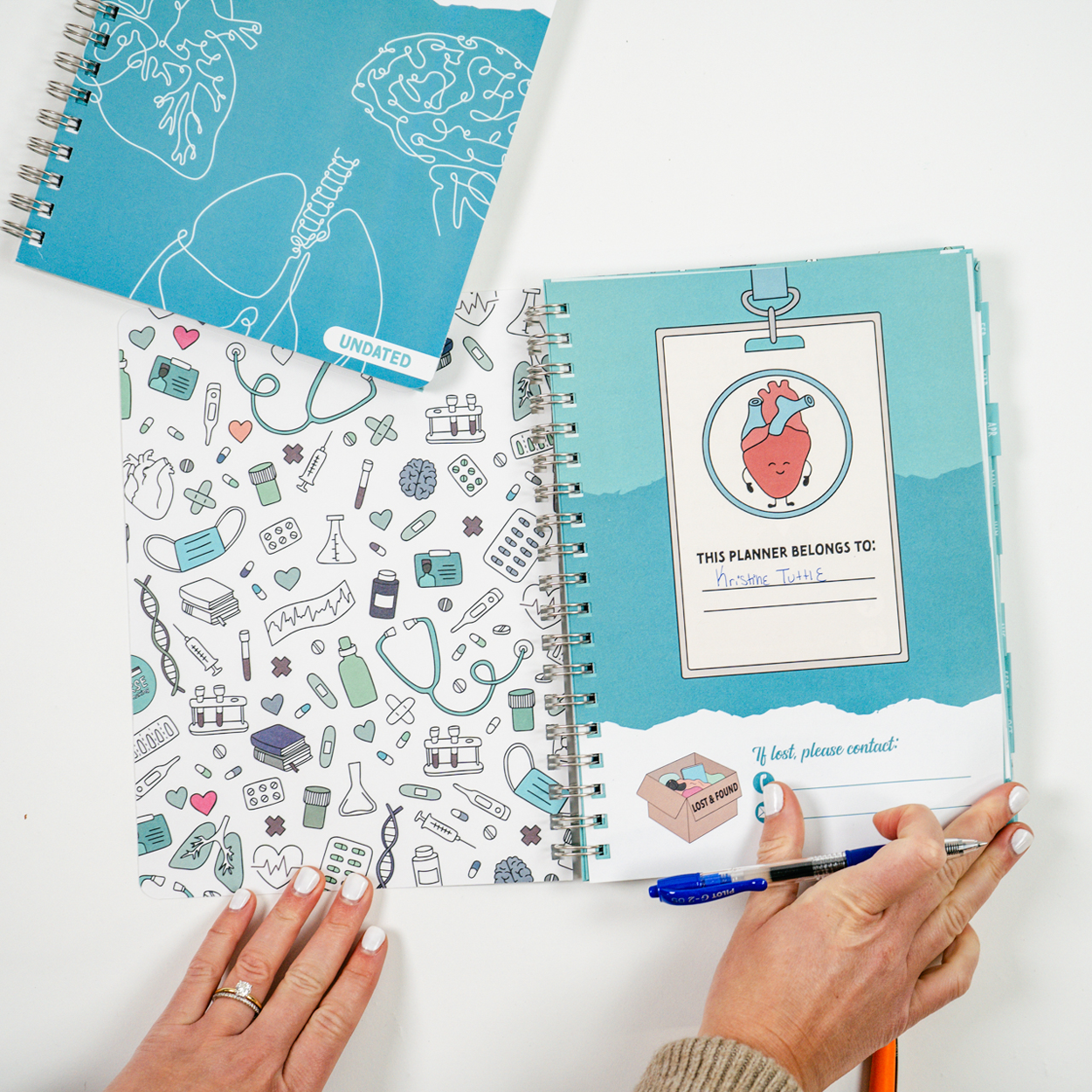 A student holds a pen while writing in a spiral-bound planner open to the name page, which features a heart illustration and a space for the owner’s name. The inside cover has a medical-themed pattern with illustrations of stethoscopes, syringes, and pill bottles. A second closed planner with a brain and lung design rests nearby.