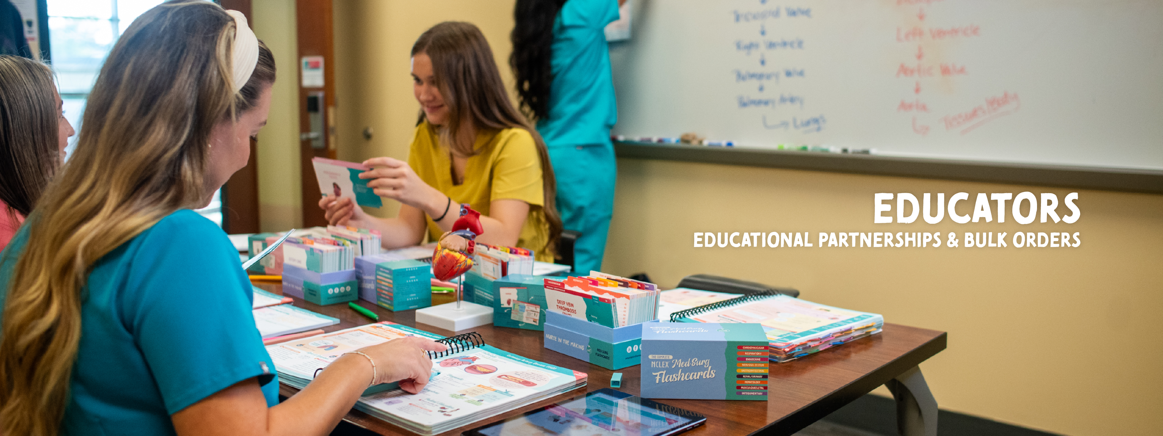 Title on the right side of the image reads “EDUCATORS — Educational Partnerships & Bulk Orders. A group of nursing students sits around a classroom table reviewing colorful study materials, including flashcards and spiral-bound guides. One student in a yellow scrub top holds a card while others look through resources. A model of a human heart sits on the table among the study tools. In the background, a whiteboard with handwritten medical notes is visible.